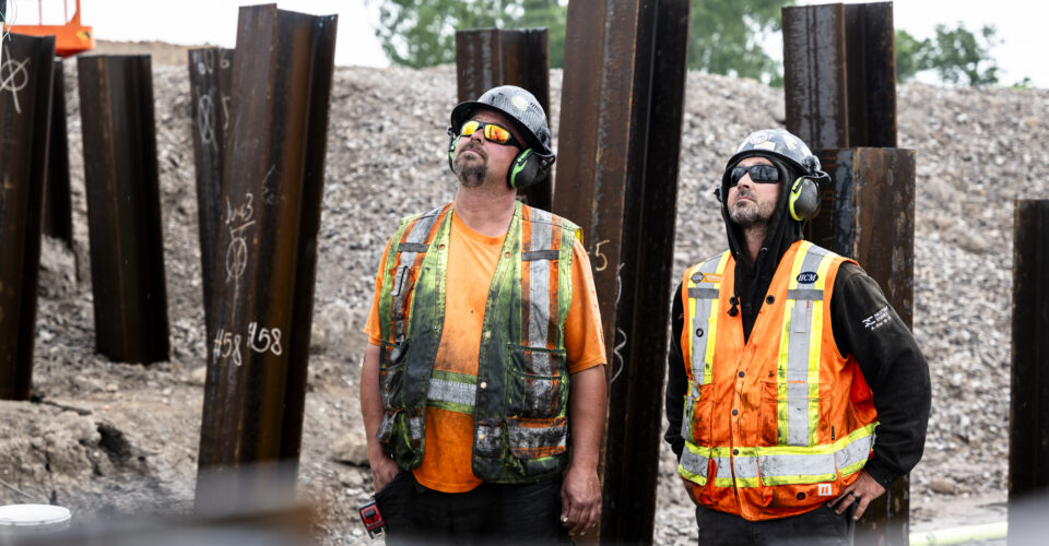 OSWCA, two employees looking up from trench, beams behind them, ear protection, 24053_BUILDWITT_1671