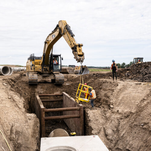 OSWCA, excavator over trench, trench box, yellow ladder, employees on either side, dirt, 24053_BUILDWITT_1841-1