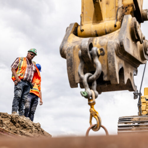 OSWCA, employee standing above trench looking down at excavator arm pulling up hooked equipment, 24053_BUILDWITT_1974