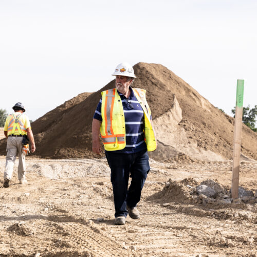 OSWCA, older employee walking in dirt, mounds and another employee in background, white hard hat, striped shirt, yellow safety vest, 24053_BUILDWITT_2510