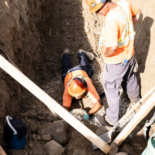 OSWCA, two employees in trench, one laying on ground, other standing next to him, pipe, wood, orange and black hard hats, vertical, 24053_BUILDWITT_2517