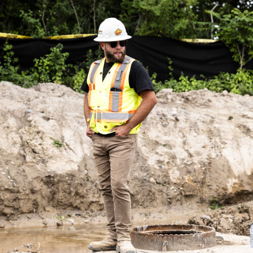 OSWCA, employee standing in dirt, white hard hat, yellow safety vest, black shirt, greenery and caution tape in background, 24053_BUILDWITT_3172