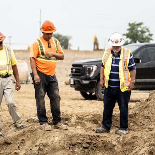 OSWCA, three employees looking down at dirt, orange hard hats, white hard hat, black truck in background, 24053_BUILDWITT_3474