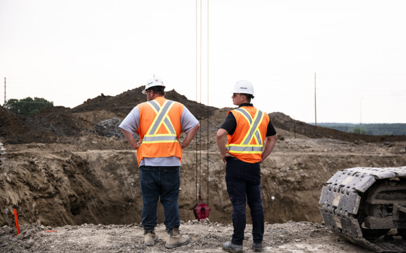 OSWCA, behind two employees looking down to trench, white hard hats, orange safety vests, 24067_BUILDWITT_2238