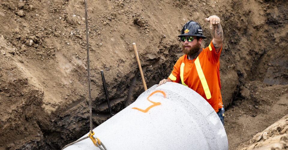 OSWCA, employee, black hard hat, orange safety shirt, hand signaling, lowering pipe, 24053_BUILDWITT_3206-1